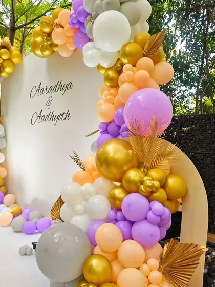 An elegant outdoor welcome baby setup for twins. The clean white backdrop is framed by a beautiful balloon garland in pastel peach, purple, and gold.