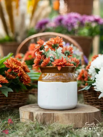A rustic detail from a boho-themed Haldi, featuring orange chrysanthemums in a traditional ceramic jar, placed on a simple wooden stump.