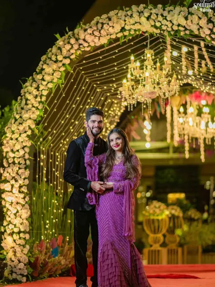 A beautiful couple poses under a floral arch illuminated with fairy lights. This makes for a perfect photo spot at a reception, combining romantic lighting with elegant decor.