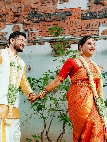 The couple holding hands, captured in a candid walking shot that highlights their traditional wedding attire and the natural greenery of the venue.