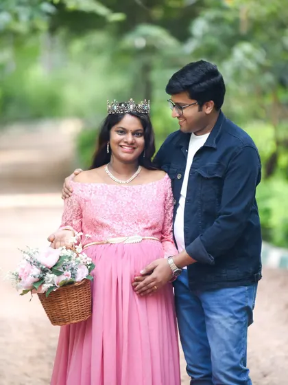 A sweet couple portrait taken on a rustic park pathway. The mom-to-be, wearing a pink dress and a tiara, holds a basket of flowers, looking radiant next to her partner.