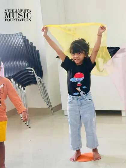 A young girl joyfully waves a yellow scarf, participating in a movement activity designed to teach musical expression. Our classes are full of color, energy, and smiles.