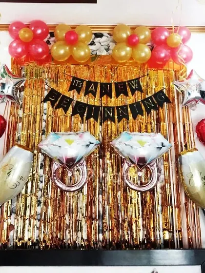 An engagement party or bridal shower backdrop. This fun setup features a gold shimmer curtain, 'It's a New Beginning' banner, and large foil balloons shaped like diamond rings and champagne glasses.