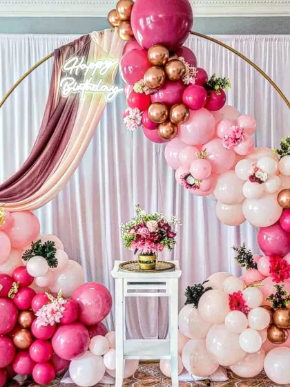 A very lush and full balloon arch in shades of pink and rose gold, with fabric draping and floral accents for a rich, textured look.