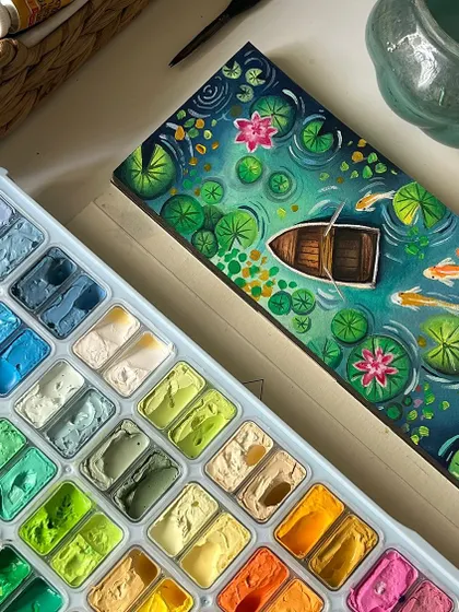 A top down view of my finished gouache lily pond painting next to my paint palette. It shows the rich texture of the gouache and the wide range of colors I used to create the scene.