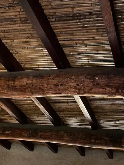 The ceiling detail of a farmhouse in Hassan, featuring heavy, reclaimed wooden beams and a layer of woven bamboo. This combination of rustic materials creates a warm, textured, and authentically vernacular space.