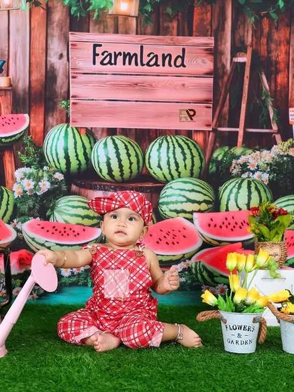 A baby girl sits in a charming "Farmland" setup, surrounded by watermelons and flowers. The rustic props and cheerful red outfit make for a unique and adorable portrait.