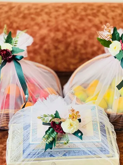Two wicker baskets filled with fresh fruits and a gift box, all part of a wedding trousseau. The wrapping uses white net and dark green ribbons for a classic and elegant presentation.