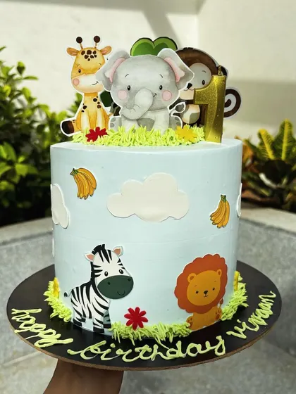 A jungle theme cake featuring a giraffe, elephant, lion, and monkey. The buttercream grass and fondant clouds add to the wild adventure.