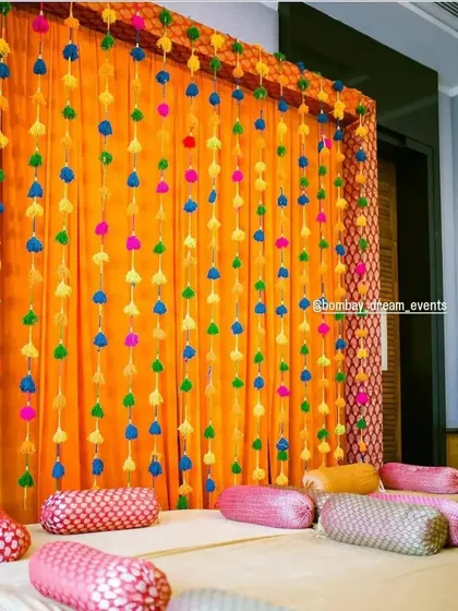 A simple yet vibrant backdrop for a home Mehndi or Dholki, using colorful pom-pom strings against a bright orange drape.