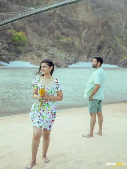 A candid-style shot of a couple on a river beach, with the woman in the foreground and the man looking towards her from the background.