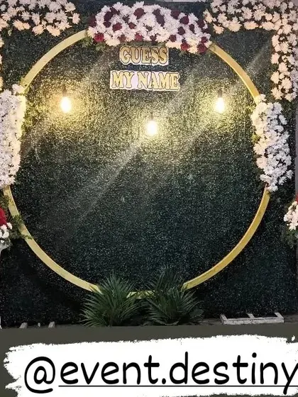 A modern naming ceremony design featuring a circular golden frame against a green wall. The frame is decorated with white and red flowers, perfect for a stylish photo booth.