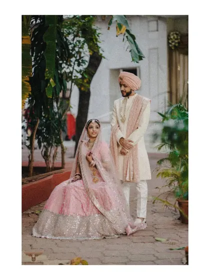 A full-length, formal portrait. The couple is posed in the courtyard of a palace, their elegant pink and white outfits complementing the regal setting.