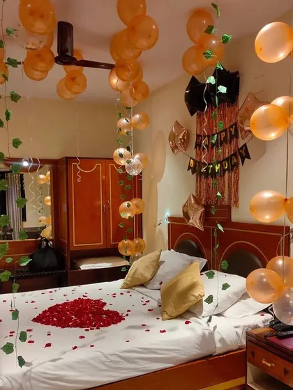 A cozy and romantic birthday room setup. The decor features peach-colored balloons, hanging ivy, a rose petal heart on the bed, and a simple "Happy Birthday" banner against a shimmer backdrop.