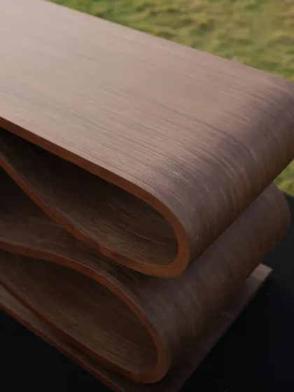 A close-up view of the SWISH bench, highlighting the smooth curves and the rich texture of the wood veneer. The layers of plywood are visible at the edges, revealing the honesty of its construction.