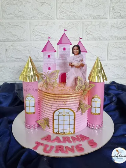 A princess castle cake fit for royalty. This design features pink turrets with golden tops, a printed image of the birthday girl, and butterfly accents.