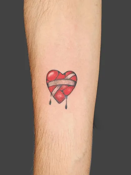 A small, broken heart tattoo held together by a bandage. This minimalist design uses a pop of red to convey deep emotion in a simple, artistic way.