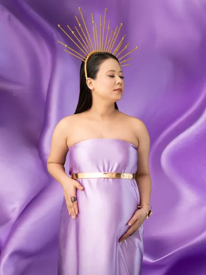A truly regal portrait. Against a backdrop of flowing purple satin, the mother-to-be wears a matching gown and a striking golden sunburst crown, embodying the divine power of creation.