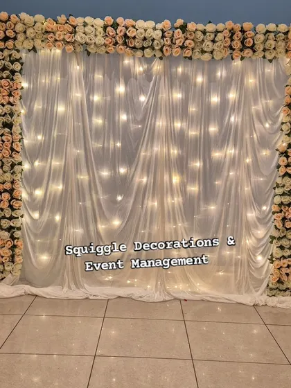 A simple yet elegant photo backdrop for any occasion. This setup features a curtain of fairy lights behind sheer white drapes, framed by a border of delicate artificial roses.