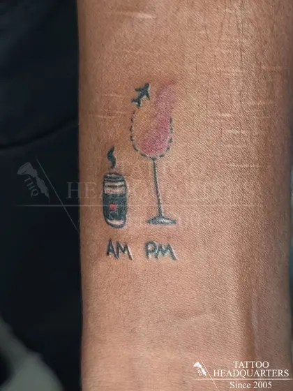 A quirky minimalist tattoo depicting a coffee cup for the "AM" and a wine glass for the "PM," symbolizing the daily routine.