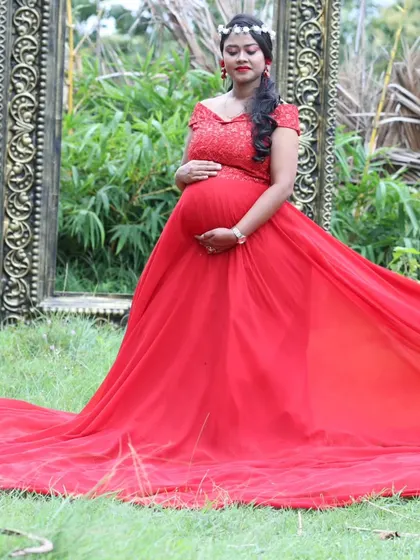 A beautiful maternity portrait in a garden, with the woman in a red trail gown posing next to an ornate golden frame prop.