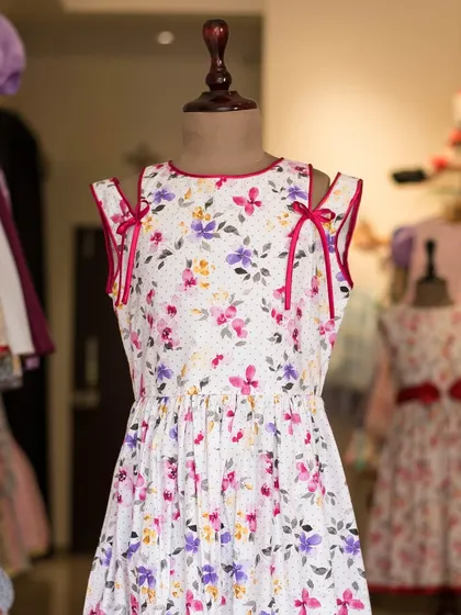 A delightful white floral dress with cold-shoulder details and pink ribbon ties. Displayed on a mannequin, you can see the light, airy feel of this perfect summer dress.