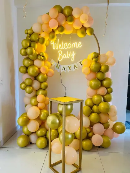 An elegant "Welcome Baby" decoration in gold and peach tones. The setup includes a balloon ring with a neon sign, a name banner, and a pedestal for the cake.