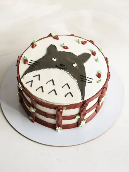A cute hand-painted cake featuring Totoro from Studio Ghibli. The design is framed with a piped border and tiny strawberries, making it a whimsical treat for an anime fan.