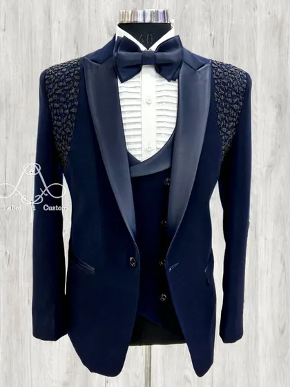 A navy blue three-piece tuxedo with a modern twist. The shoulders feature a textured, beaded pattern, adding a unique detail to the classic satin shawl collar and pleated formal shirt.