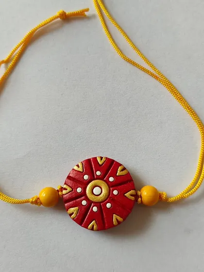 This rakhi features a traditional red and gold design on a terracotta base with a bright yellow thread. It's a classic and auspicious color combination.