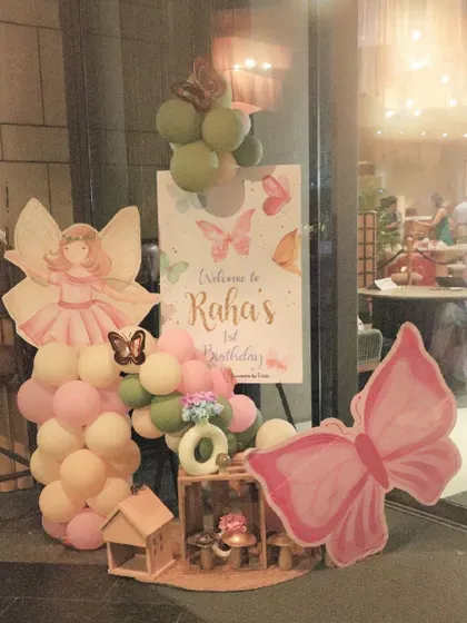 A charming welcome sign for Raha's first birthday. This setup combines a personalized easel sign with fairy and butterfly cutouts and small balloon clusters, creating a sweet first impression for guests.