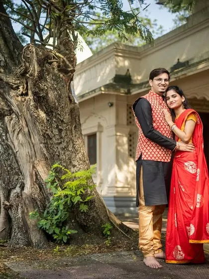 A classic portrait of a couple in traditional Indian outfits, posing by an old, majestic tree on historic grounds.