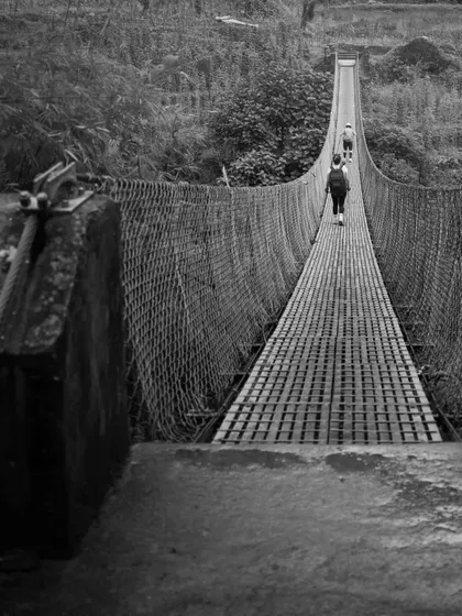A black and white shot of trekkers crossing a long suspension bridge, highlighting the dramatic and adventurous elements of the journey to Annapurna Base Camp.
