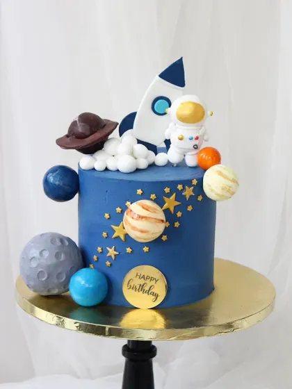 An out-of-this-world space-themed cake for a little astronaut. This deep blue cake is decorated with edible planets, golden stars, and a fondant astronaut and rocket ship ready for liftoff.