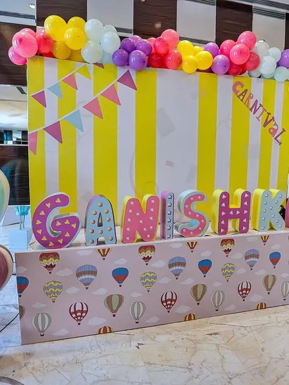 A personalized carnival name display for "Ganishka." The setup features large, colorfully decorated letters, a hot-air-balloon-themed table skirt, and a vibrant yellow-and-white striped backdrop topped with a balloon garland.