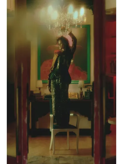 A scene of daring expression from "Love in Future Past." The composition, with the model standing on a chair under a chandelier, is meant to feel spontaneous and defiant, breaking from the quiet nostalgia of earlier chapters.