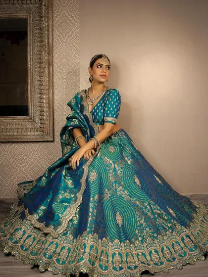 A model posing in the beautiful teal banarasi lehenga. The look is completed with a traditional choker and heavy earrings, showcasing a complete rental package for any grand occasion.