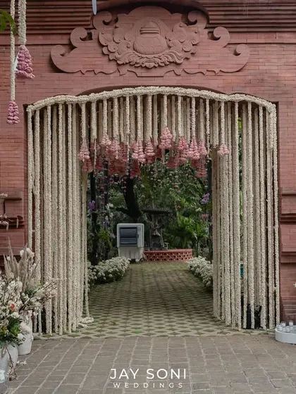 A beautiful fusion of traditional and rustic elements. This wedding entrance features a gate draped in tuberose garlands against a terracotta brick wall, flanked by arrangements of pampas grass and white florals in large urns.