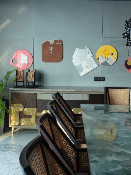 The dining room features a signature sideboard from Furgonomics with 'Thathera' brass work. The wall is adorned with a colorful collection of artworks by Tanmoy Samanta, creating a vibrant and artistic dining experience.