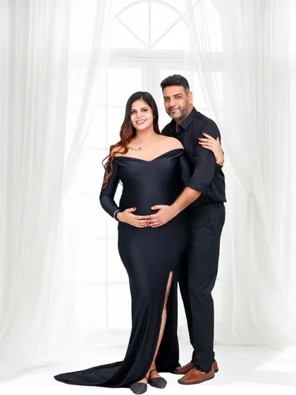 A classic, full-length studio portrait of an expecting couple. The woman wears a sleek black gown, and the bright, airy background provides a timeless and elegant feel.