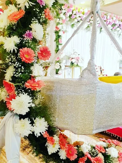 An elegant cradle decoration for a modern naming ceremony. The silver cradle is framed by a lush arrangement of fresh flowers, including pink gerberas and white chrysanthemums, creating a soft and celebratory look.