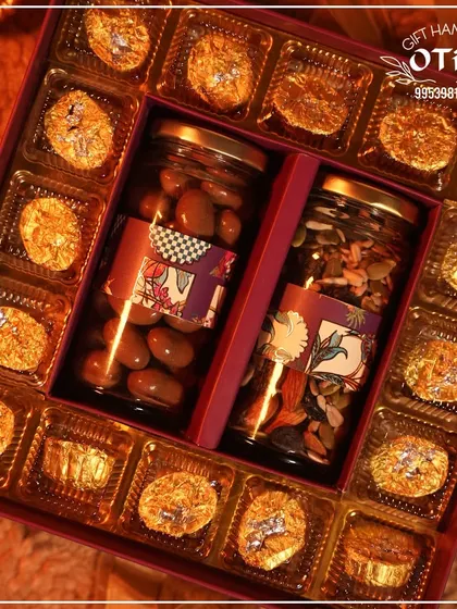 An overhead view of a luxurious Diwali chocolate box. It features two central jars of chocolate almonds and mixed seeds, surrounded by gold-wrapped premium chocolates.
