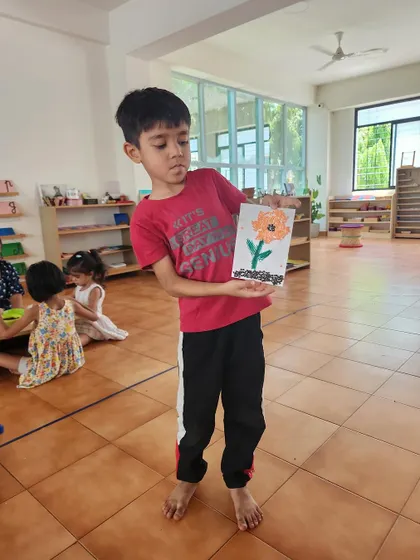 A young artist proudly presents his finished piece, a flower made with seeds. This project combines drawing, gluing, and working with natural materials.