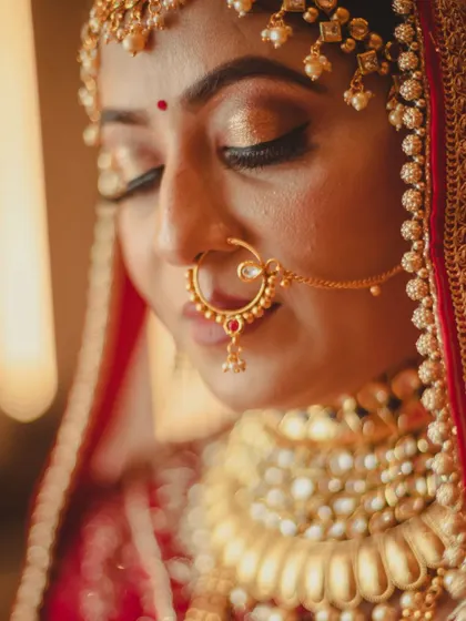 A close-up portrait focusing on the bride's delicate features and stunning jewelry. The soft focus on the background draws all attention to her shimmering gold eyeshadow and the intricate design of her nath.