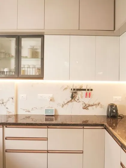 A U-shaped kitchen with glossy off-white cabinets and a brown granite countertop. The design includes rose gold profile handles, adding a subtle touch of luxury and warmth to the modern aesthetic.
