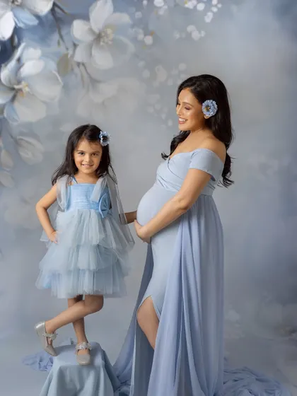 A sweet mother-daughter moment during a maternity session. The mom-to-be looks lovingly at her daughter, who is striking a cute pose, both dressed in beautiful matching blue dresses.