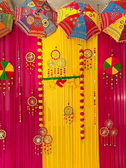 A vibrant Haldi ceremony backdrop. The decor uses colorful drapes in pink and yellow, decorated with traditional Indian umbrellas and floral hangings.