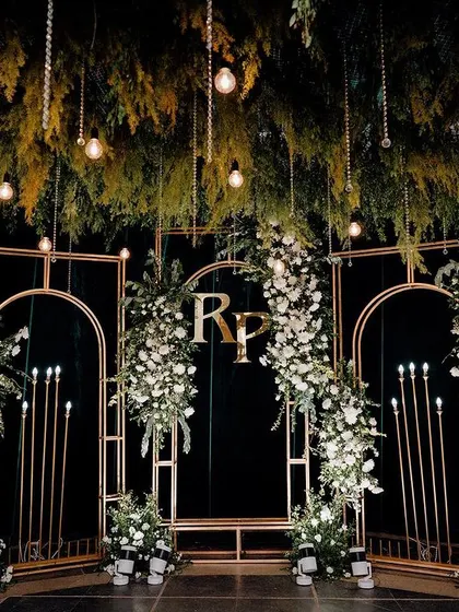 A close-up of the Sangeet stage backdrop, showing the couple's monogram, gold arches, and lush white floral arrangements against a dark green curtain.
