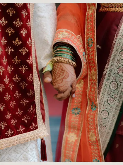 A close-up detail shot of the couple holding hands, showing the bride's green bangles and mehndi against the groom's traditional attire.