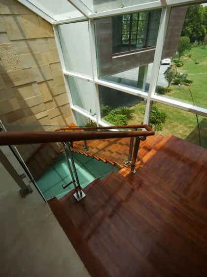 This staircase in the Sachdeva Farmhouse is enclosed within expansive glass panels, bathing the space in natural light and framing views of the garden. The warm wooden treads contrast with the textured Indian stone walls.